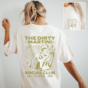 May include: A white t-shirt with a gold graphic print of a woman's face and the text "THE DIRTY MARTINI 24/7 365 IT'S TINI TIME! SOCIAL CLUB Est. THE FILTHIER THE BETTER 1951"