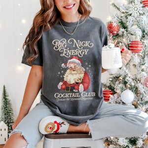 Puede incluir: Camiseta gris oscuro con un gr&aacute;fico de Pap&aacute; Noel gui&ntilde;ando un ojo, con un traje y gorro rojos. El texto "Big Nick Energy" y "Cocktail Club Fueled by Espresso Martinis!" est&aacute;n impresos en la camiseta. La persona sostiene una taza blanca con un dise&ntilde;o de &aacute;rbol de Navidad.