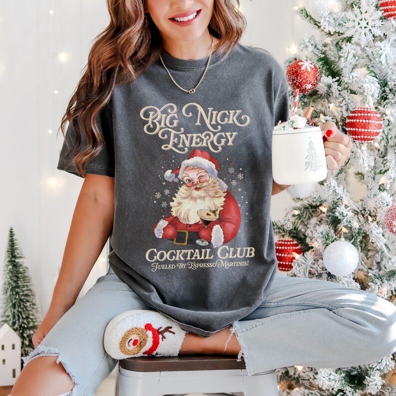Puede incluir: Camiseta gris oscuro con un gr&aacute;fico de Pap&aacute; Noel gui&ntilde;ando un ojo, con un traje y gorro rojos. El texto "Big Nick Energy" y "Cocktail Club Fueled by Espresso Martinis!" est&aacute;n impresos en la camiseta. La persona sostiene una taza blanca con un dise&ntilde;o de &aacute;rbol de Navidad.