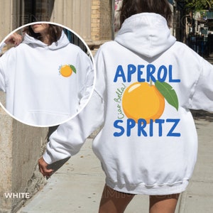 May include: White hoodie with a front pocket and a hood. The back of the hoodie features the words "APEROL SPRITZ" in blue, with a large orange graphic and a green leaf. The phrase "Ciao Bella!" is also printed on the back.