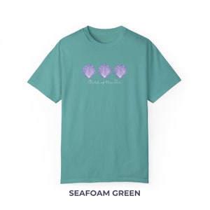 Pu&ograve; includere: T-shirt verde acqua con una grafica di tre conchiglie viola e il testo "Child of the Sea".