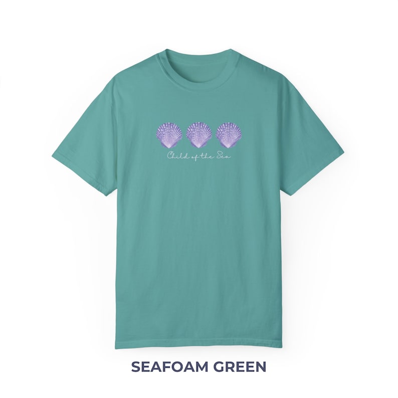 Pu&ograve; includere: T-shirt verde acqua con una grafica di tre conchiglie viola e il testo "Child of the Sea".