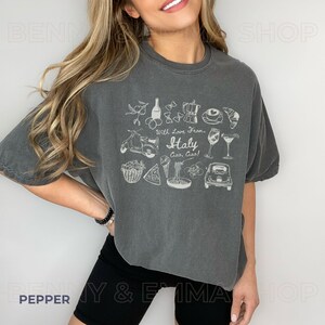 May include: A grey cropped t-shirt with a white graphic design featuring Italian icons such as a Vespa scooter, pizza, pasta, and wine. The text on the shirt reads "With love from... Italy Ciao, Ciao!"