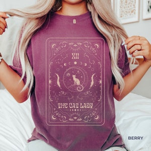 Pode incluir: Uma camiseta roxa com um gr&aacute;fico de uma carta de tar&ocirc; com um gato sentado em uma lua crescente. A carta &eacute; cercada por estrelas e outros s&iacute;mbolos celestiais. O texto "THE CAT LADY" &eacute; impresso abaixo da imagem.