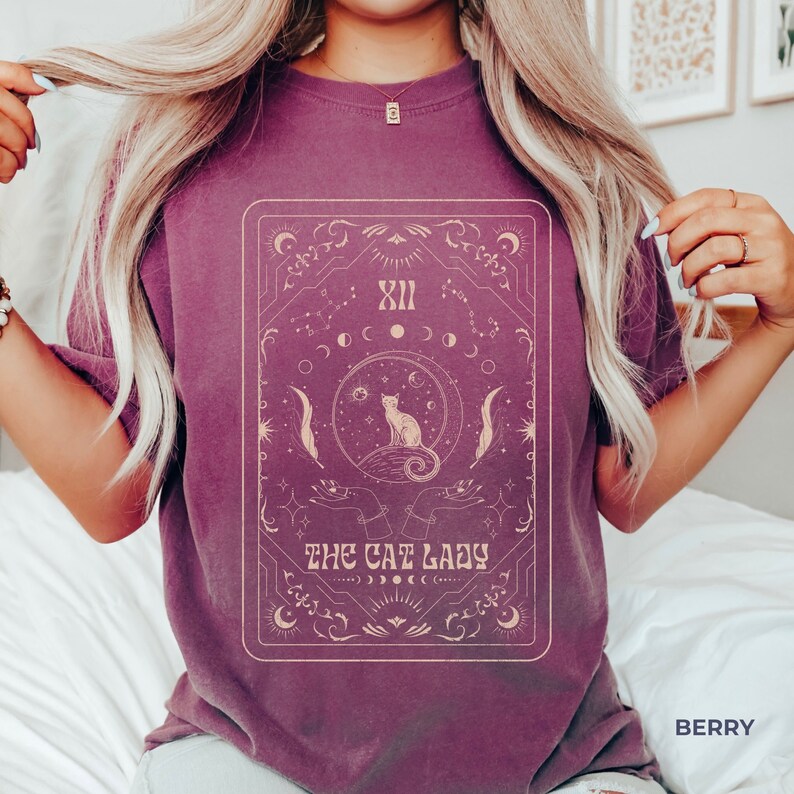 Pode incluir: Uma camiseta roxa com um gr&aacute;fico de uma carta de tar&ocirc; com um gato sentado em uma lua crescente. A carta &eacute; cercada por estrelas e outros s&iacute;mbolos celestiais. O texto "THE CAT LADY" &eacute; impresso abaixo da imagem.