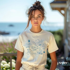 May include: A woman wearing a white t-shirt with a blue graphic design featuring a lighthouse, whale, and other nautical elements. The graphic also includes the text "With love from Martha's Vineyard 2022". The t-shirt is labeled "IVORY".