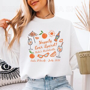 May include: White sweatshirt with an orange and black graphic design. The design includes illustrations of cocktails, oranges, sunglasses, and the text "Happily Ever Aperol! Katie's Bachelorette, Costa D'Amalfi - July, 2025."
