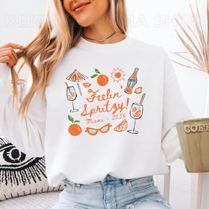 May include: White sweatshirt with a graphic design of a cocktail glass, orange slices, sunglasses, a sun, an umbrella, and a bottle. The text on the sweatshirt says "Feelin' Spritzy! Miami - 2025".