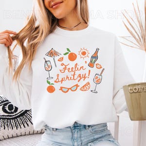 May include: White sweatshirt with a graphic design of a cocktail glass, orange slices, sunglasses, and the text "Feelin' Spritzy!"