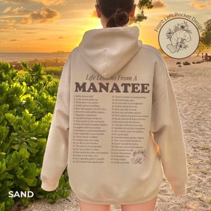K&ouml;nnte beinhalten: Ein beiger Hoodie mit einem braunen Grafikdruck eines Manatis und einer Liste von Lebenslektionen eines Manatis. Der Text enth&auml;lt S&auml;tze wie "Chill is the new thrill" und "Let the current guide you".
