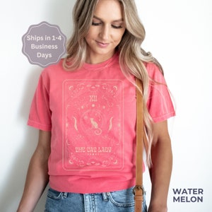 Pode incluir: Uma camiseta rosa com um design de carta de tar&ocirc; de estilo vintage que apresenta um gato e o texto "The Cat Lady".