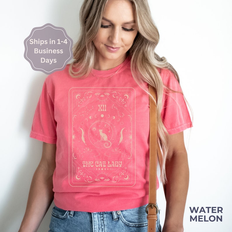 Pode incluir: Uma camiseta rosa com um design de carta de tar&ocirc; de estilo vintage que apresenta um gato e o texto "The Cat Lady".