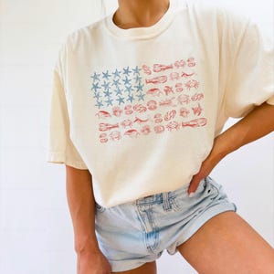 May include: Cream-colored t-shirt featuring a unique graphic of the American flag. The flag design replaces stars with blue starfish and stripes with red sea creatures. The shirt is paired with light blue denim shorts.