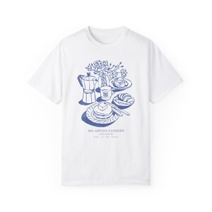 May include: White t-shirt featuring a blue illustration of a breakfast scene, including a coffee pot, a cup of coffee, a plate of pancakes, and a croissant. The text "Bon Appetit Patisserie Cafe & Bistro Paris 5th 75005 France" is printed below the illustration.