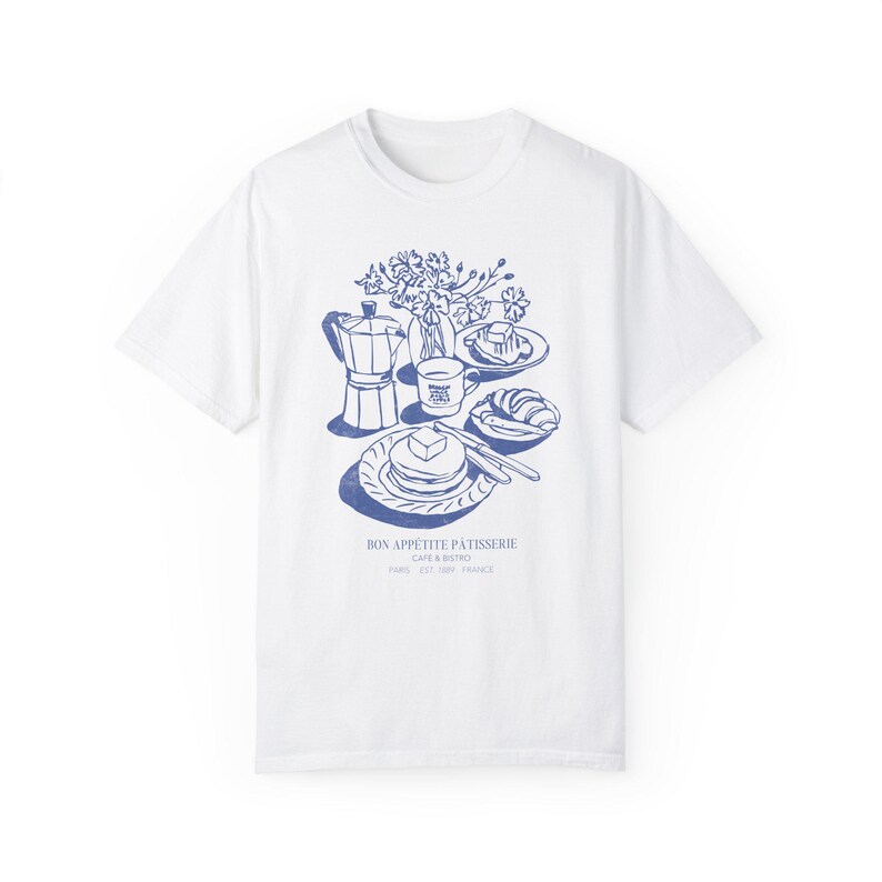 May include: White t-shirt featuring a blue illustration of a breakfast scene, including a coffee pot, a cup of coffee, a plate of pancakes, and a croissant. The text "Bon Appetit Patisserie Cafe & Bistro Paris 5th 75005 France" is printed below the illustration.