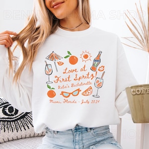 May include: White sweatshirt with a graphic design featuring a cocktail, sunglasses, oranges, and the text "Love at First Spritz! Katie's Bachelorette Miami, Florida - July 2024".