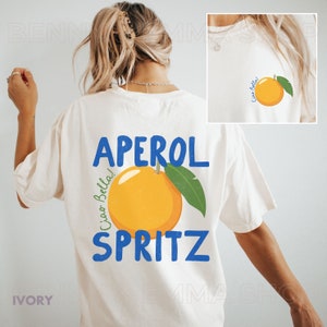 May include: White t-shirt with a blue and orange graphic that says "Aperol Spritz" and "Ciao Bella!"  The graphic features an orange slice with green leaves.