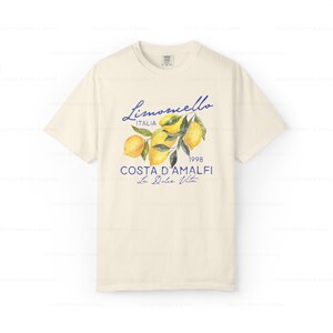 Puede incluir: Una camiseta de color crema con un estampado gr&aacute;fico azul y amarillo de limones en una rama. El texto "Limoncello Italia 1998 Costa D'Amalfi La Dolce Vita" est&aacute; impreso en la camiseta.