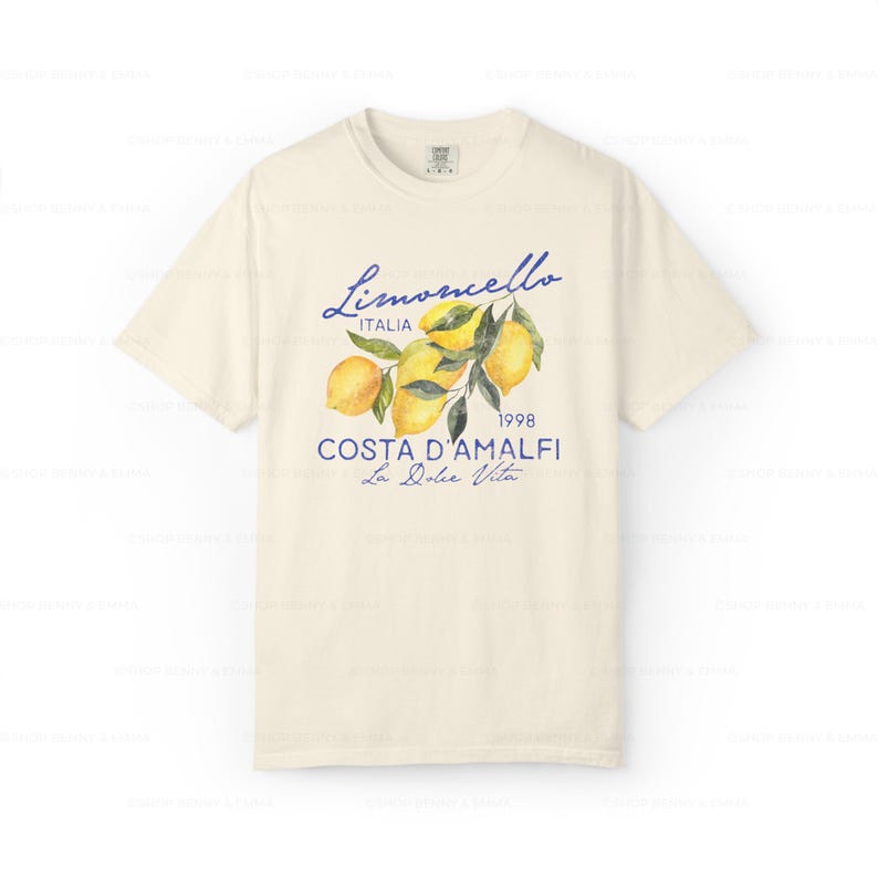 Puede incluir: Una camiseta de color crema con un estampado gr&aacute;fico azul y amarillo de limones en una rama. El texto "Limoncello Italia 1998 Costa D'Amalfi La Dolce Vita" est&aacute; impreso en la camiseta.