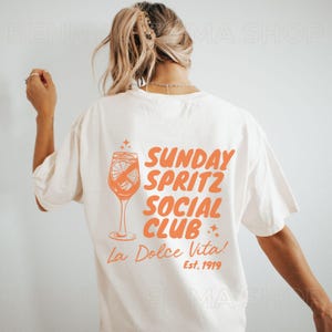 May include: White t-shirt with an orange graphic of a glass of spritz with the text "Sunday Spritz Social Club La Dolce Vita! Est. 1919"