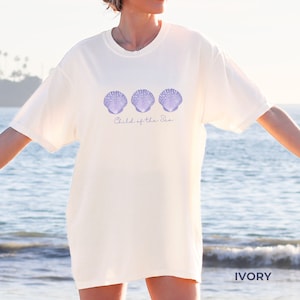 Pu&ograve; includere: Una maglietta bianca con una grafica di tre conchiglie viola e il testo "Child of the Sea". La maglietta &egrave; mostrata su un modello in piedi su una spiaggia.