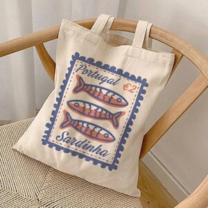 May include: A white canvas tote bag with a blue and red stamp design featuring the text "Portugal €2 Sardinha" and three stylized fish.