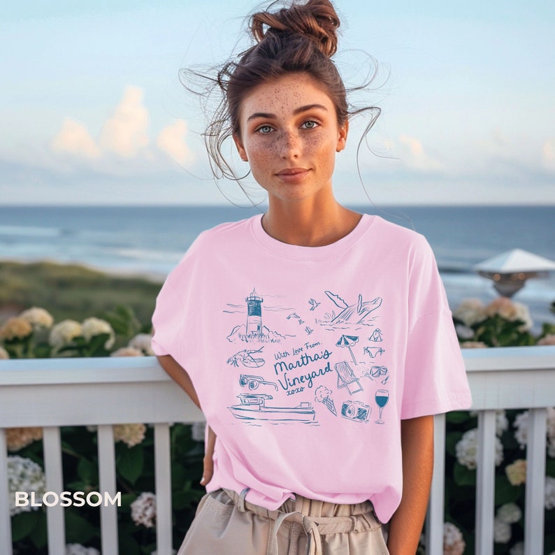 May include: A pink t-shirt with a blue graphic design featuring a lighthouse, whale, beach umbrella, and other summer-themed imagery. The text on the shirt reads "With love from Martha's Vineyard 2022".