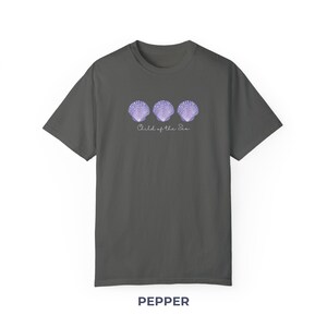 Pu&ograve; includere: Una maglietta grigia scura con un design di tre conchiglie viola e il testo "Child of the Sea".