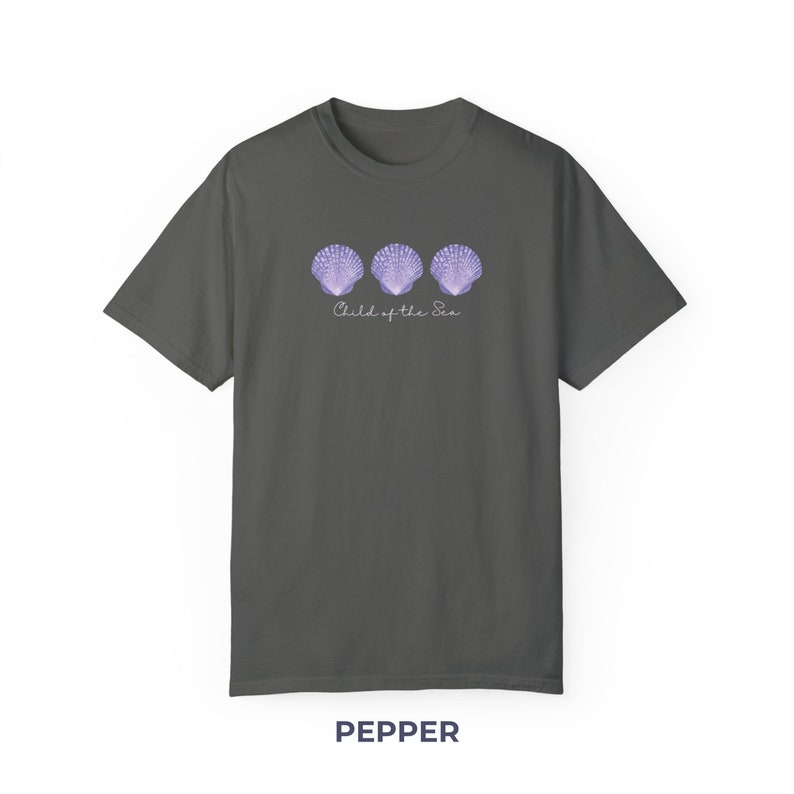 Pu&ograve; includere: Una maglietta grigia scura con un design di tre conchiglie viola e il testo "Child of the Sea".
