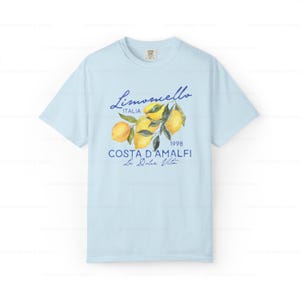 Puede incluir: Camiseta azul claro con un gr&aacute;fico de limones en una rama, el texto "Limoncello Italia 1998 Costa D'Amalfi La Dolce Vita" est&aacute; impreso en la camiseta.