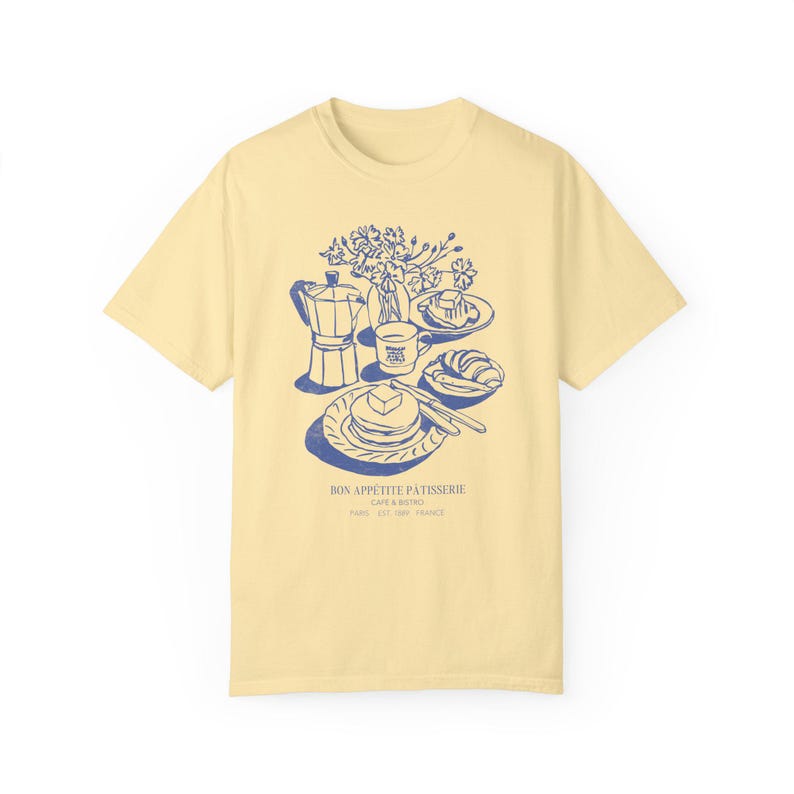 May include: A yellow t-shirt featuring a blue illustration of a breakfast scene. The illustration includes a coffee pot, a cup of coffee, a plate of pancakes, a croissant, and a vase of flowers. The text "Bon Appetit Patisserie Cafe & Bistro Paris Est. 1981 France" is printed below the illustration.