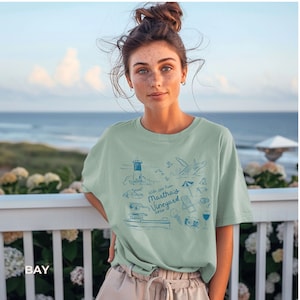 May include: A woman wearing a light green t-shirt with a blue graphic design of a lighthouse, lobster, and other beach-themed imagery. The graphic includes the text "With love from Martha's Vineyard 2023".