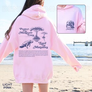 May include: Light pink hoodie with a graphic print of a manatee swimming in a mangrove forest. The text "Protect the Mangrove Trees" and "Save the Manatees" is printed on the back of the hoodie.