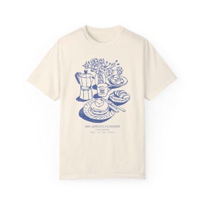 May include: A cream-coloured t-shirt featuring a blue illustration of a breakfast scene. The illustration includes a coffee pot, a cup of coffee, a plate of pancakes, a croissant, and a vase of flowers. The text "Bon Appetit Patisserie Cafe & Bistro Paris 8th 75008 France" is printed below the illustration.