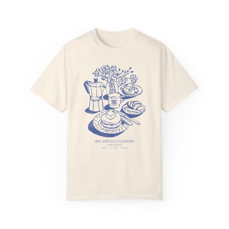 May include: A cream-coloured t-shirt featuring a blue illustration of a breakfast scene. The illustration includes a coffee pot, a cup of coffee, a plate of pancakes, a croissant, and a vase of flowers. The text "Bon Appetit Patisserie Cafe & Bistro Paris 8th 75008 France" is printed below the illustration.