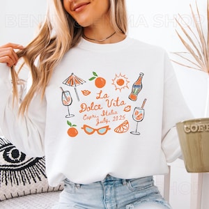 May include: White sweatshirt with a graphic design of a sun, an umbrella, a glass of orange juice, sunglasses, oranges, and the text "La Dolce Vita Capri, Italia July, 2025".