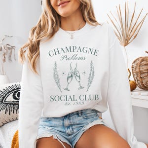 May include: White sweatshirt with a green graphic design that says "Champagne Problems Social Club Est. 1989". The graphic features two champagne glasses clinking together.