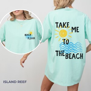May include: A light blue t-shirt with the text "Take me to the beach" on the back. The text is in black with a yellow sun and blue waves. The front of the shirt has the text "Beach Please" in black with a yellow sun and blue waves.