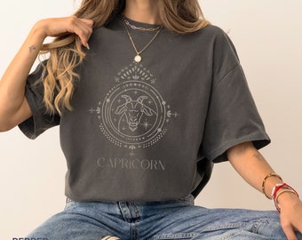 Könnte beinhalten: Ein dunkelgraues T-Shirt mit einem Grafikdruck eines Ziegenkopfes in einem Kreis, unter dem das Wort "CAPRICORN" steht. Die Grafik ist in hellgrau gehalten.