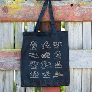 May include: Black tote bag with a white outline of different types of pasta, including tagliatelle, farfalle, ravioli, rigatoni, pappardelle, cavatelli, gnocchi, maccheroni, conchigliette, orecchiette, and penne. The bag has the text "NY & EMMA" printed on the bottom.