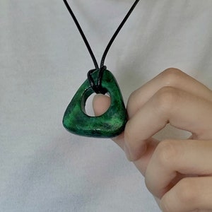 May include: A green pendant necklace with a triangular shape and a circular cutout. The pendant is a mottled green color and is suspended from a black cord. The necklace is being held by a person.