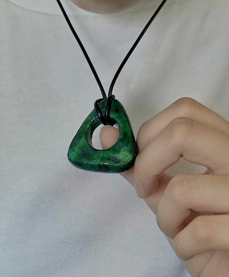 May include: A green pendant necklace with a triangular shape and a circular cutout. The pendant is a mottled green color and is suspended from a black cord. The necklace is being held by a person.