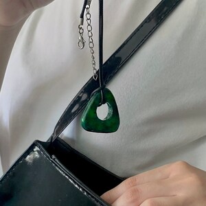 May include: A dark green triangular pendant necklace with a circular hole in the center. The pendant hangs from a black cord with a silver-colored chain and clasp. The necklace is displayed against a white shirt and a black purse.