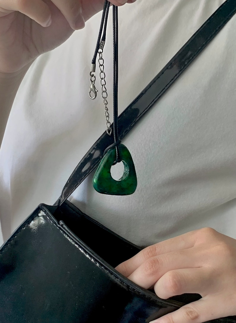 May include: A dark green triangular pendant necklace with a circular hole in the center. The pendant hangs from a black cord with a silver-colored chain and clasp. The necklace is displayed against a white shirt and a black purse.