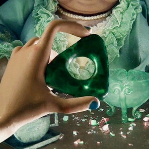 May include: A dark green planchette with a circular opening is held up. A matching green bowl with three legs and a decorative face is nearby. The scene has a vintage aesthetic, with a person in a green outfit and a pearl necklace in the background.