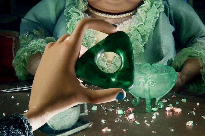 May include: A dark green planchette with a circular opening is held up. A matching green bowl with three legs and a decorative face is nearby. The scene has a vintage aesthetic, with a person in a green outfit and a pearl necklace in the background.
