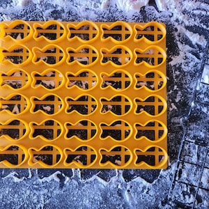 May include: A yellow plastic cookie cutter with a fish shape. The cutter is on a black surface with white powder on it.