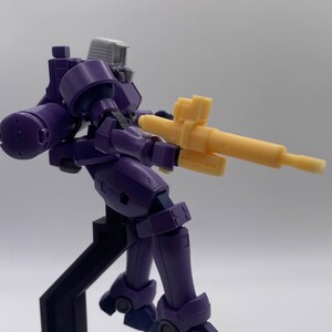 May include: A purple and gray robot action figure holding a yellow gun. The robot is posed in a dynamic action pose with one leg raised. The figure is standing on a black stand.
