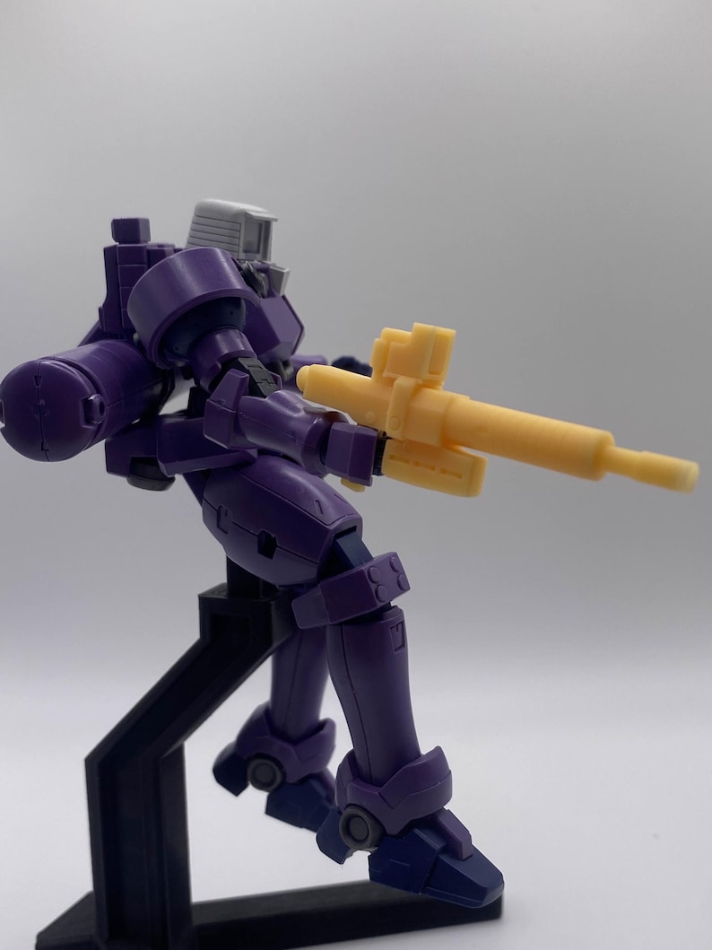 May include: A purple and gray robot action figure holding a yellow gun. The robot is posed in a dynamic action pose with one leg raised. The figure is standing on a black stand.