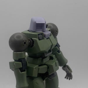 以下が含まれることがあります： コクピット付きの緑と灰色のプラスチック製のロボットモデルキット。ロボットは大きく丸い肩と大きく丸い頭をしています。